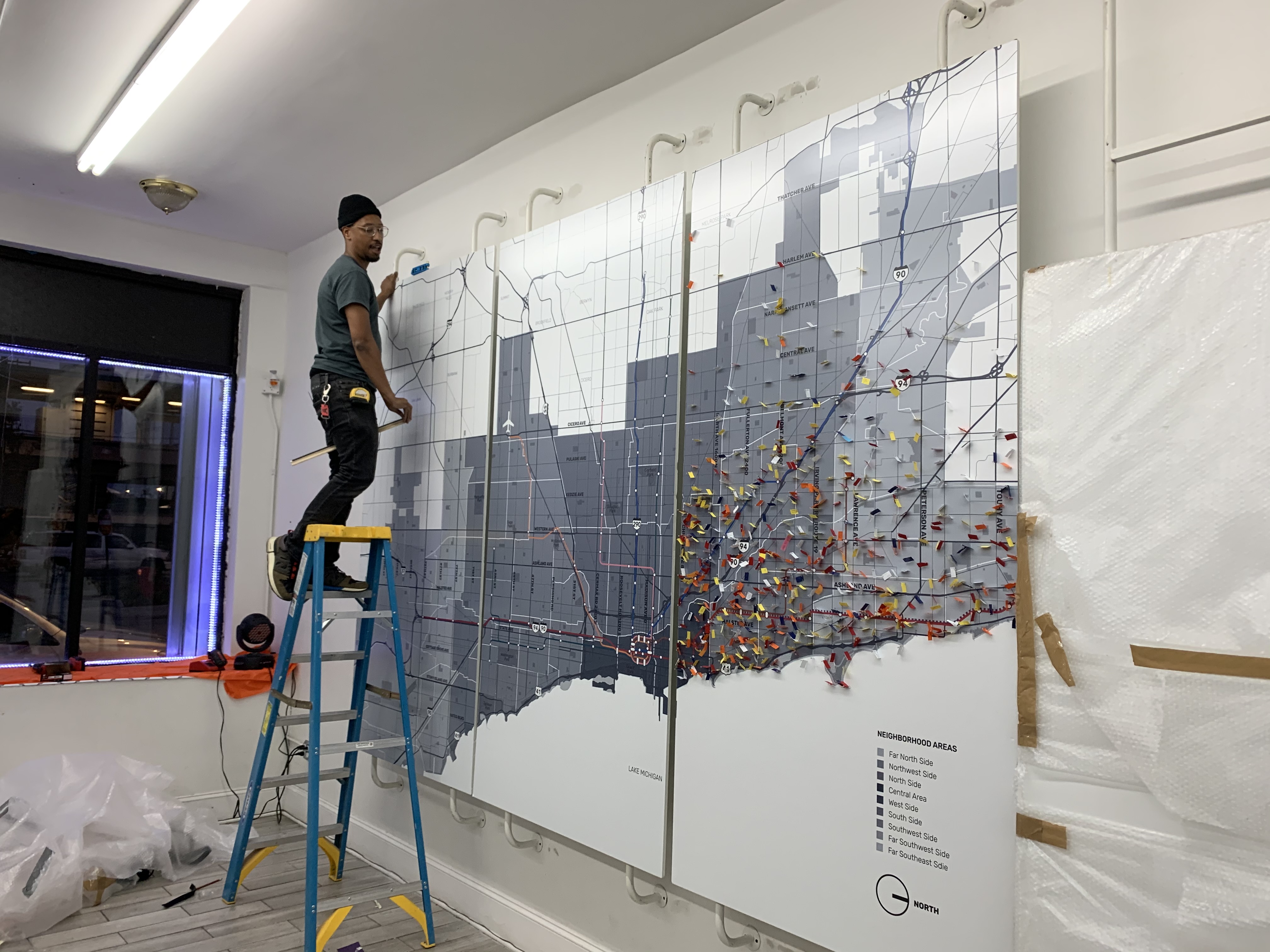 A Black man on a ladder reaches to the back of a 8' by 12' map of Chicago affixed to the wall by tubing that has flags of various colors pinned onto it.